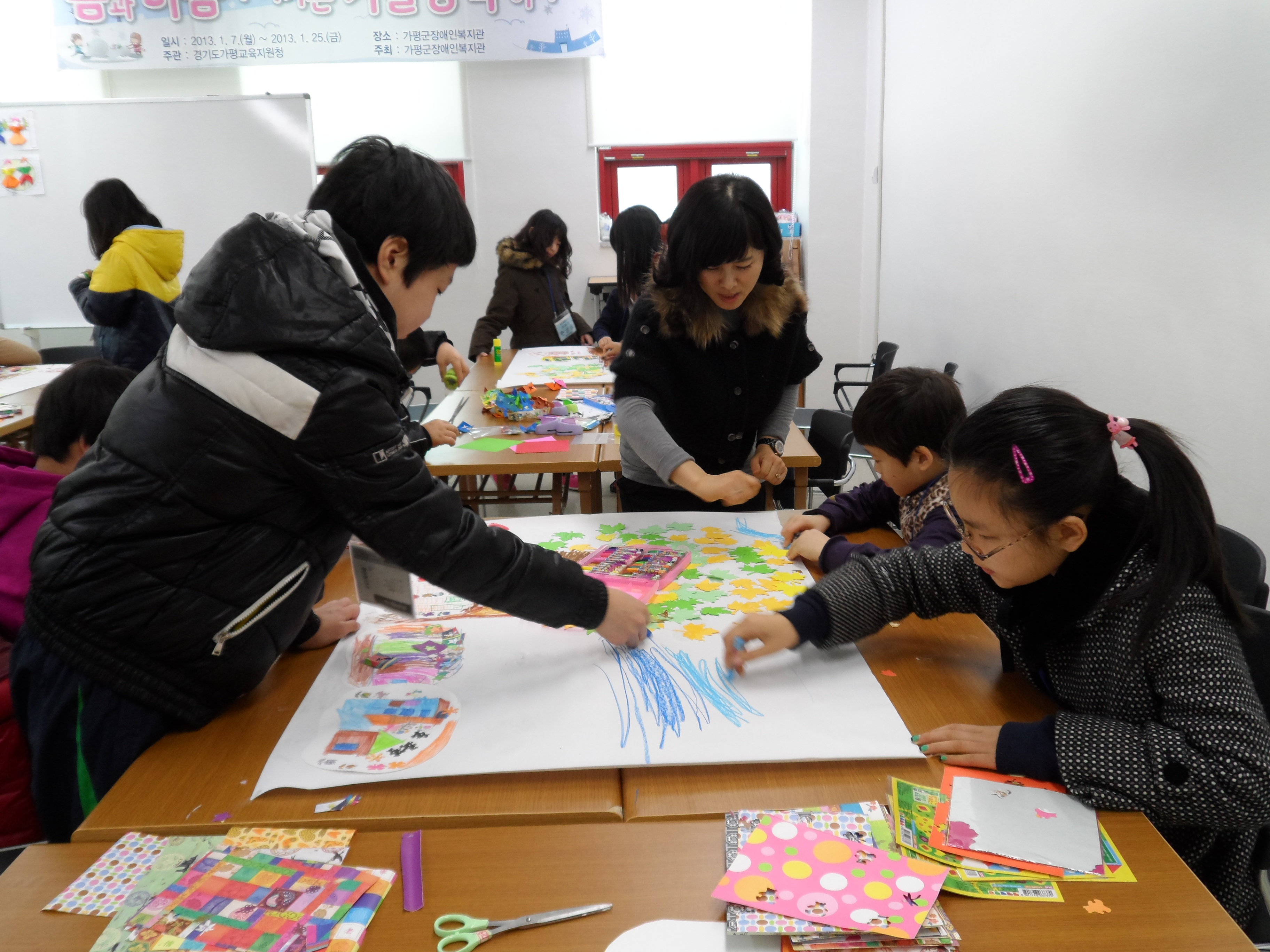 장애 학생들이 여자선생님의 설명을 들으며 큰 도화지 위에 여러가지 색깔의 크레파스를 이용해 색칠을 하고 있는 모습